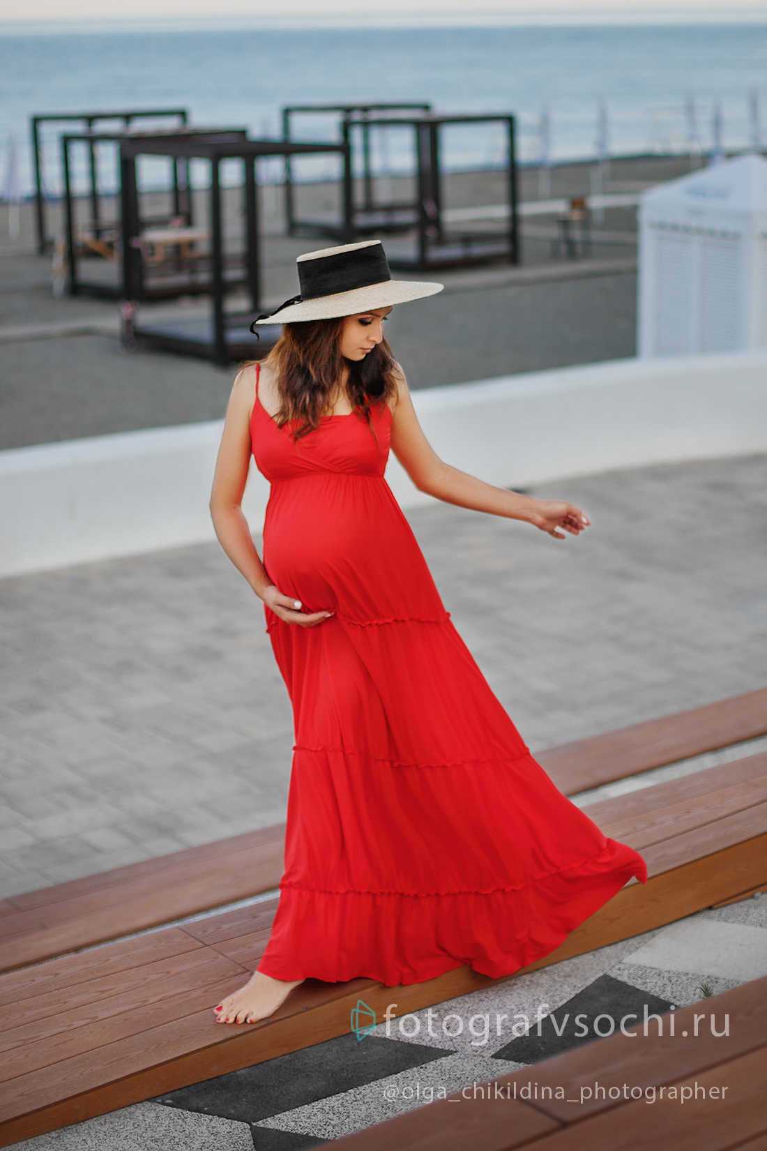 Беременная девушка в красном платье и шляпке прогуливается у моря