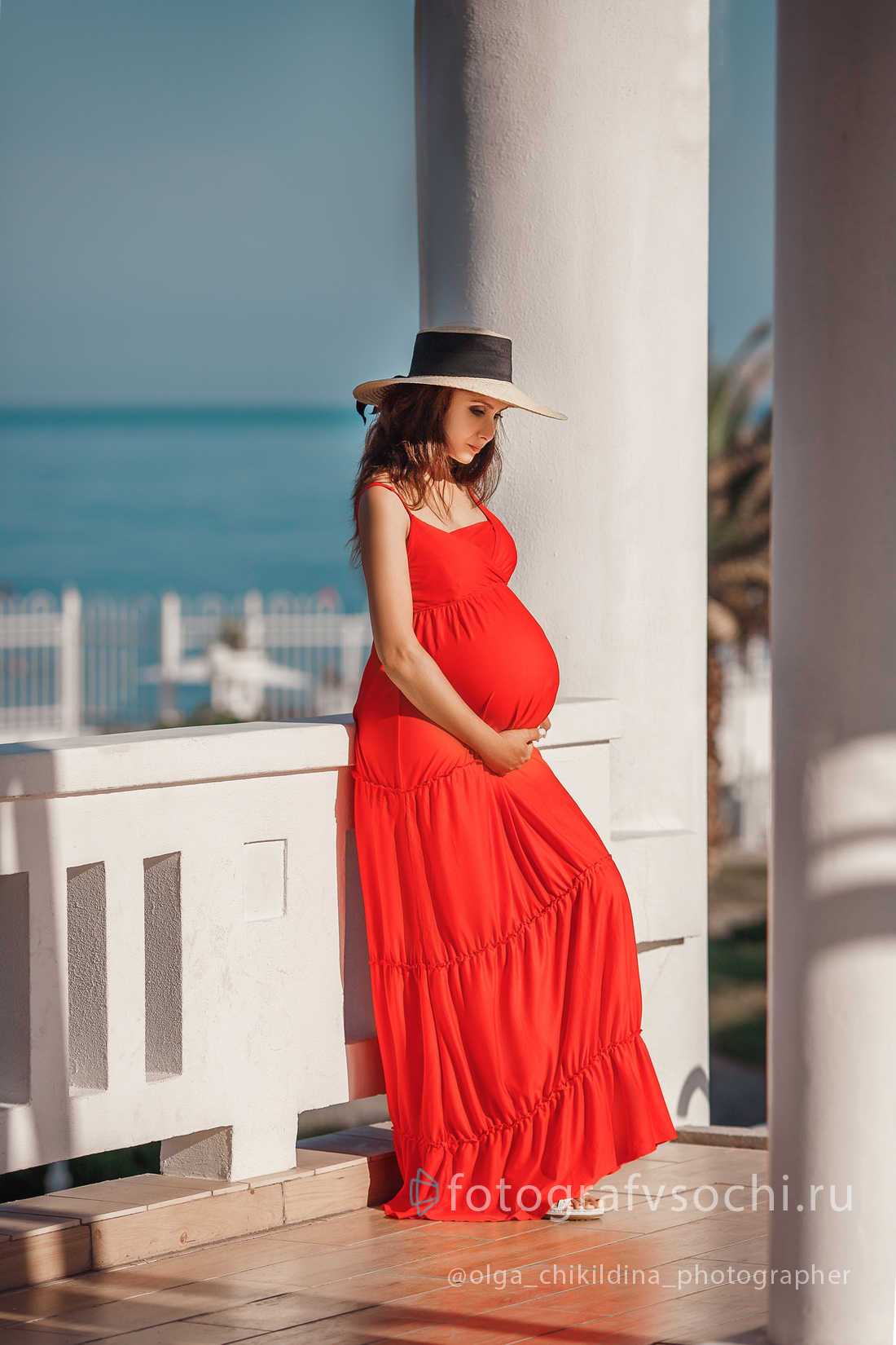 Беременная девушка в шляпке и красном платье на фоне белой колонны и моря