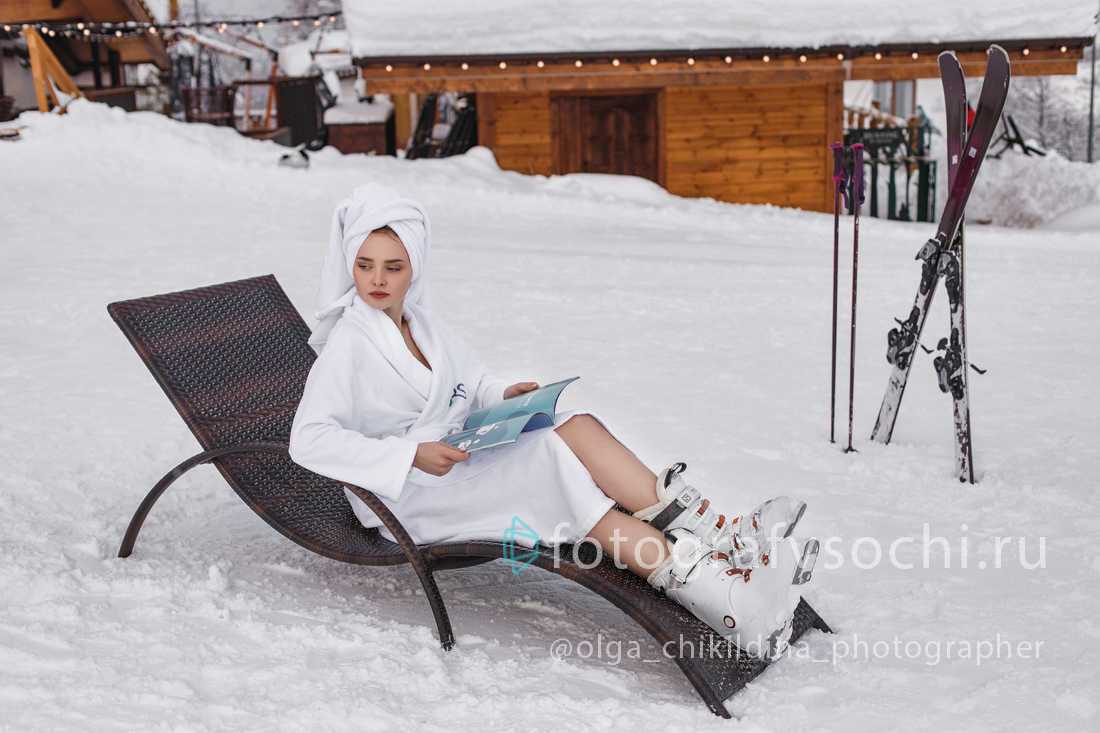 Лыжница в халате на лежаке с журналом, вокруг нее лежит снег