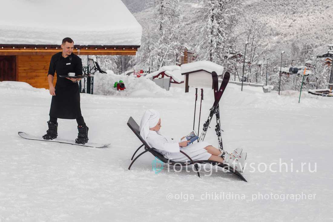 Официант на сноуборде подвозит кофе девушке в халате лежащей на лежаке прямо на заснеженной улице
