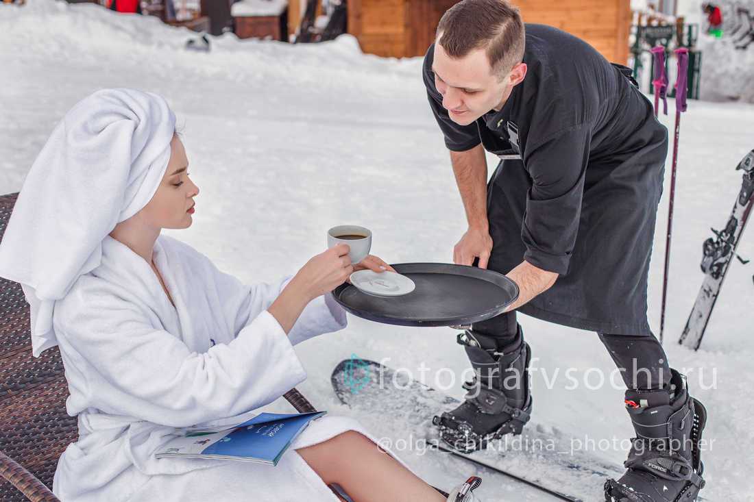 Официант на сноуборде подносит кофе девушке в халате и полотенцем на головке сидящей на лежаке прямо на заснеженной улице