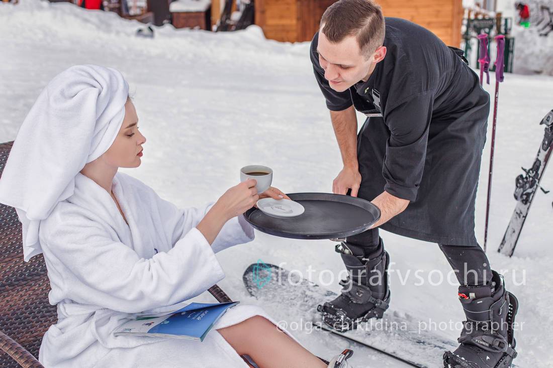 Официант на сноуборде подносит кофе девушке в халате и полотенцем на головке сидящей на лежаке прямо на заснеженной улице